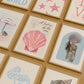 Set of 10 coastal watercolor prints in pink and blue: "Wild Like the Sea" beside a spiral shell illustration, "Stay Wild Ocean Child" in bold blue lettering, "Sea La Vie" in pink stamped typography, a mermaid lounging in a martini glass, pink starfish and seashell illustrations, a woman in a shell headdress, a beach umbrella watercolor, and "Meet me by the sea" in postcard-style script.