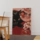 Vertical photographic print of a black cat with amber eyes resting beside two pink martini cocktails garnished with tiny orange pumpkins, on a deep red surface with a woman's hand holding one glass stem.