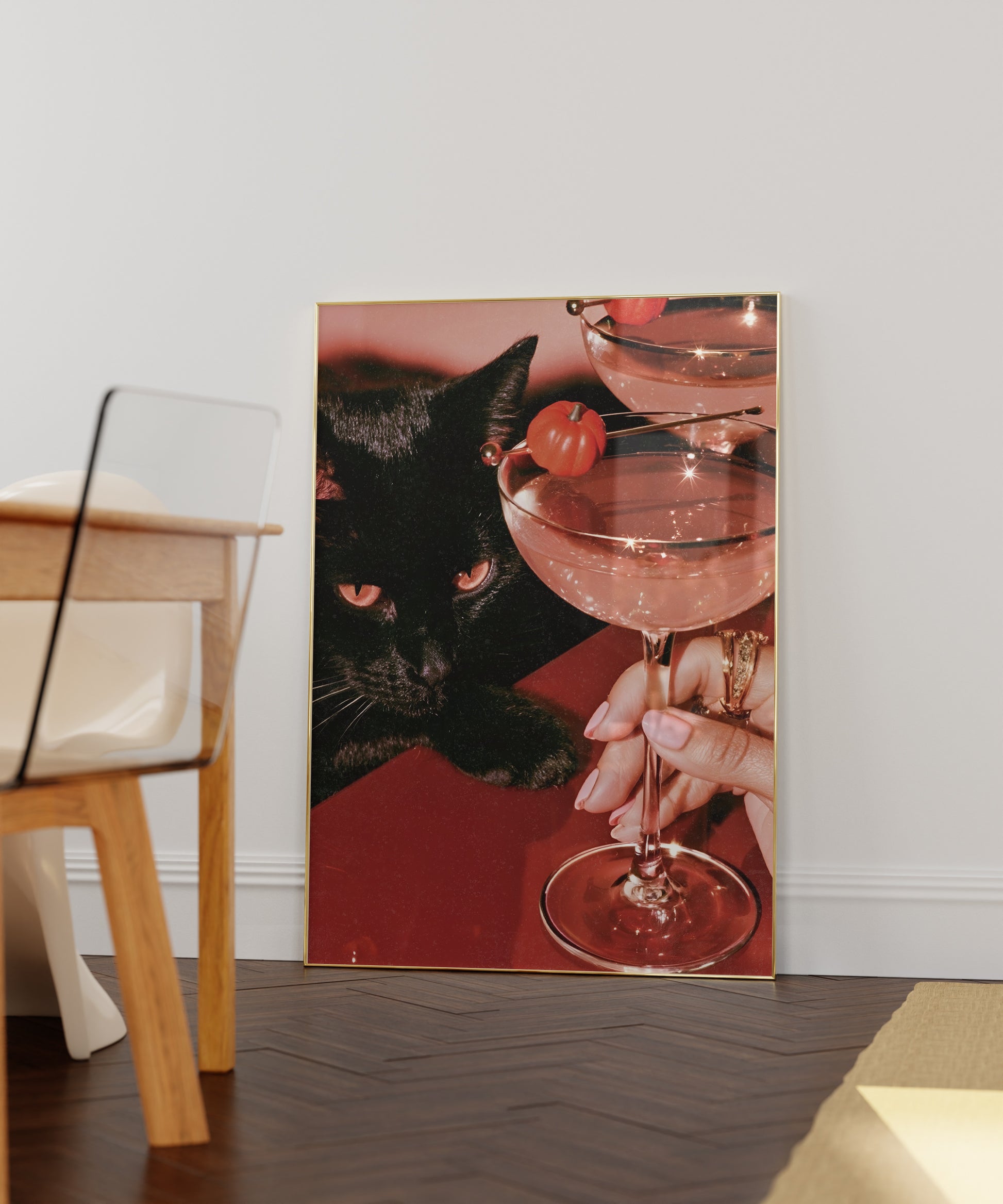 Vertical photographic print of a black cat with amber eyes resting beside two pink martini cocktails garnished with tiny orange pumpkins, on a deep red surface with a woman's hand holding one glass stem.