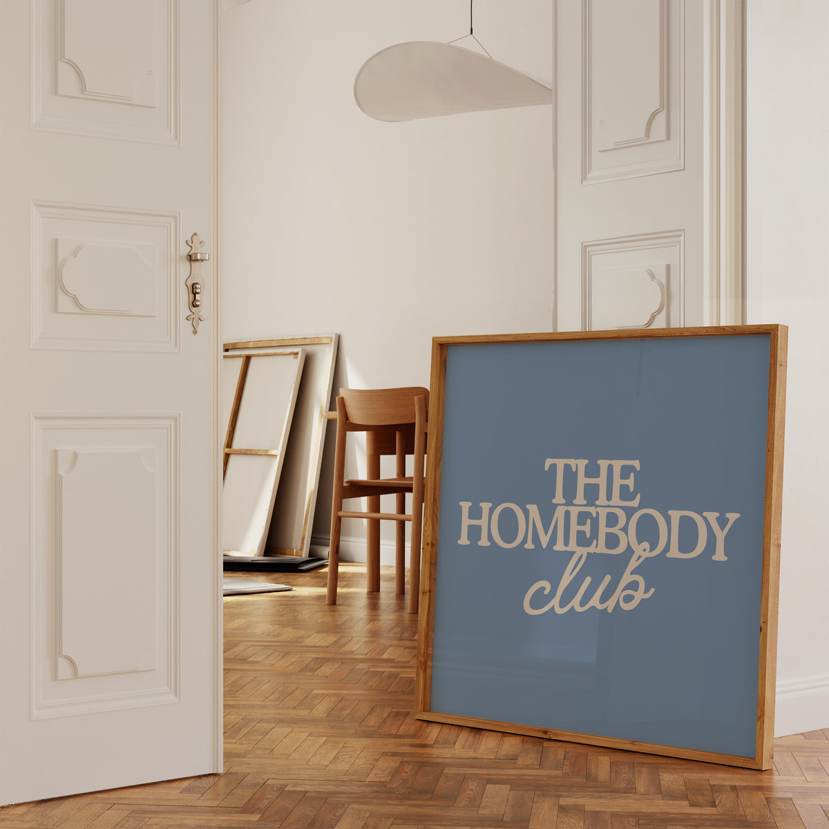 Square digital print with a dusty blue background featuring "THE HOMEBODY" in bold cream serif type stacked above "club" in cream cursive script, centered typography layout.