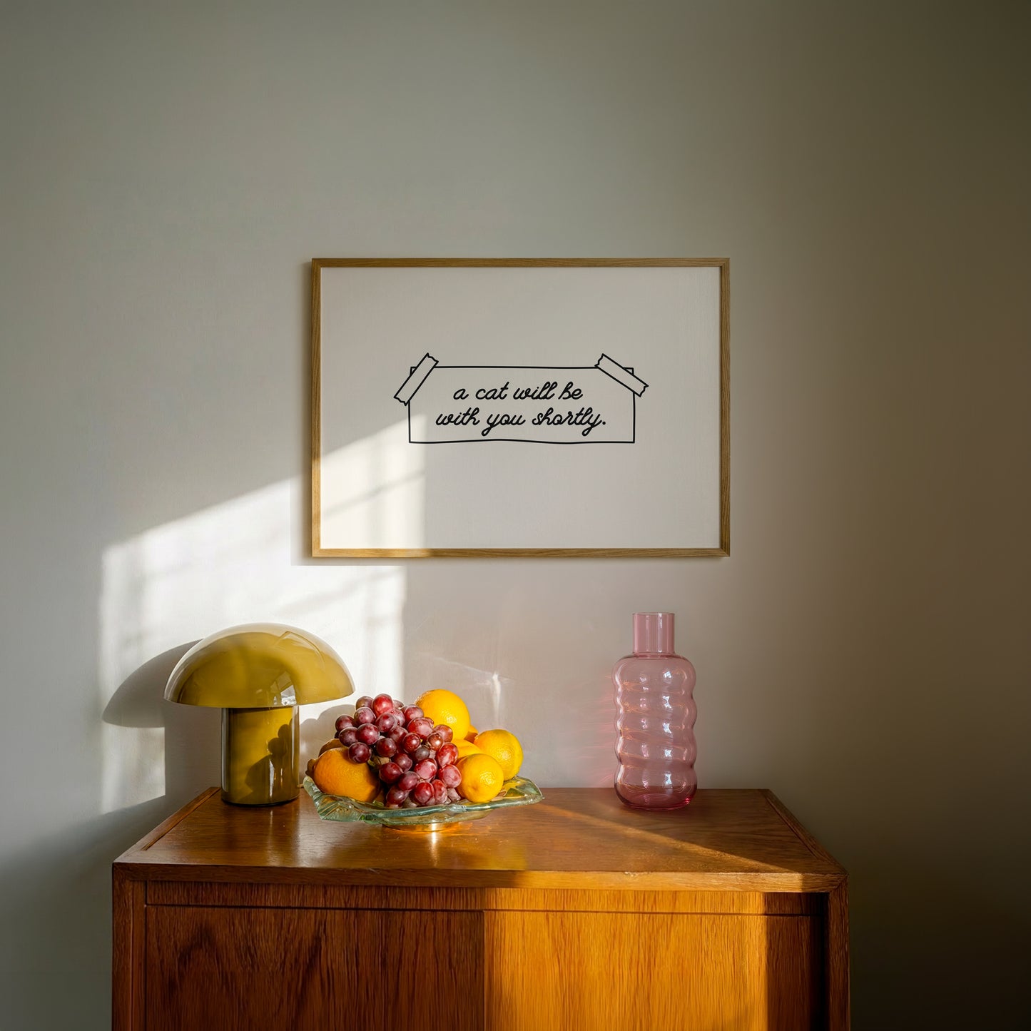 Horizontal wall print on a white background with a hand-drawn taped note in cursive script reading a cat will be with you shortly
