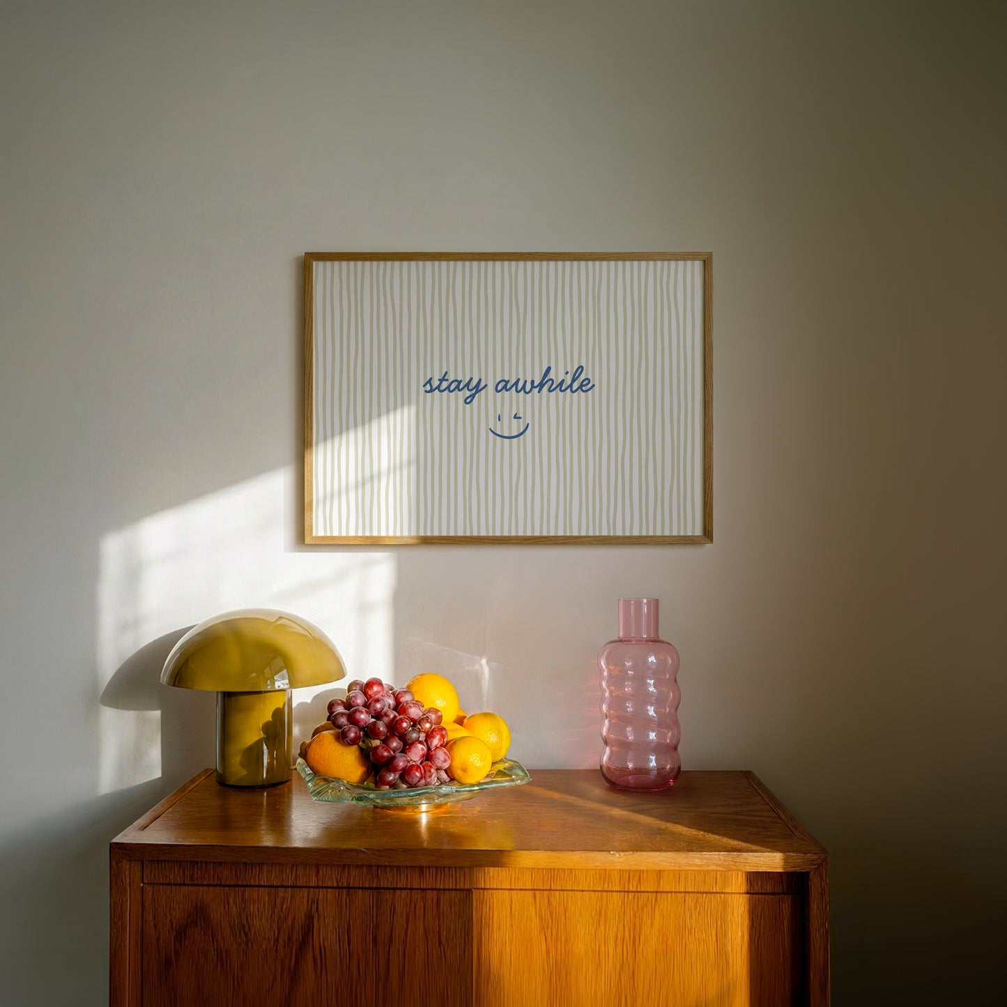 Horizontal print with thin hand-painted tan and cream vertical stripes, the quote "stay awhile" in navy cursive script, and a small hand-drawn smiley face beneath it.