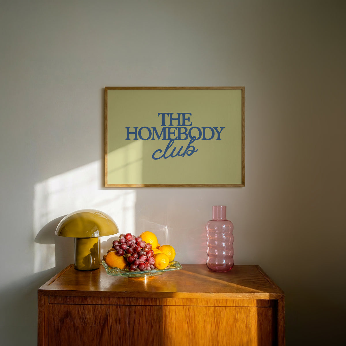 Rectangular landscape digital print with an olive-green background featuring "THE HOMEBODY" in bold blue serif type stacked above "club" in blue cursive script, centered typography layout.