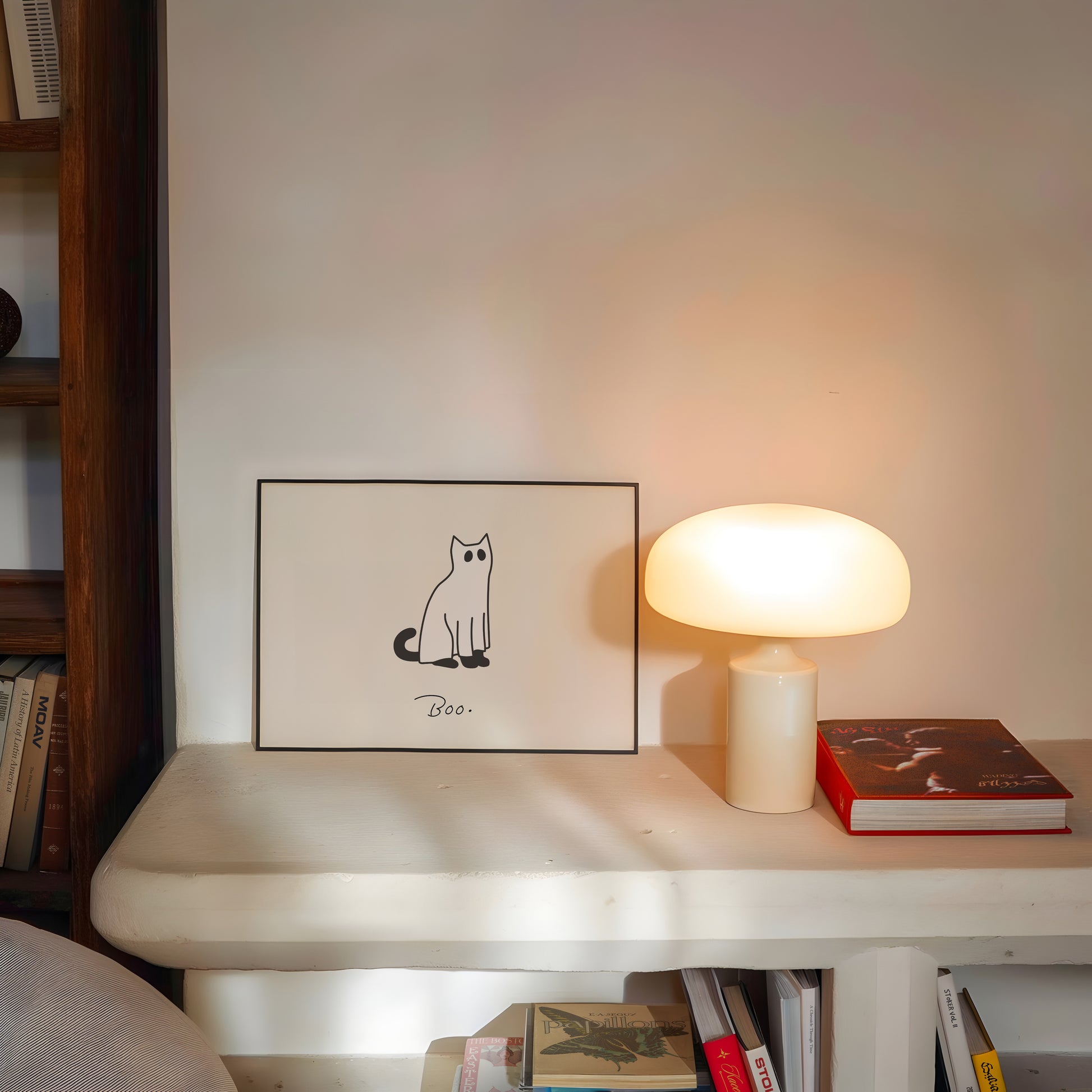 Horizontal minimalist print of a black cat wearing a white ghost sheet costume with cut-out eyes, sitting beside the handwritten word "Boo." on a cream background.