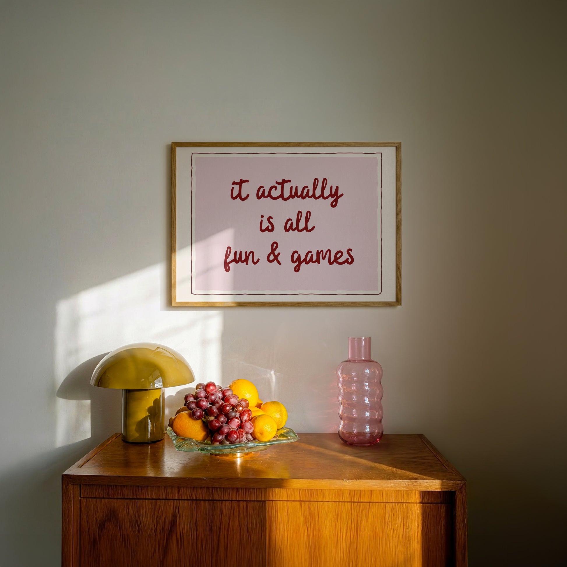 Horizontal wall print with a soft pink centre panel and white border with dark red scalloped outline, bold dark red cursive script reading it actually is all fun & games
