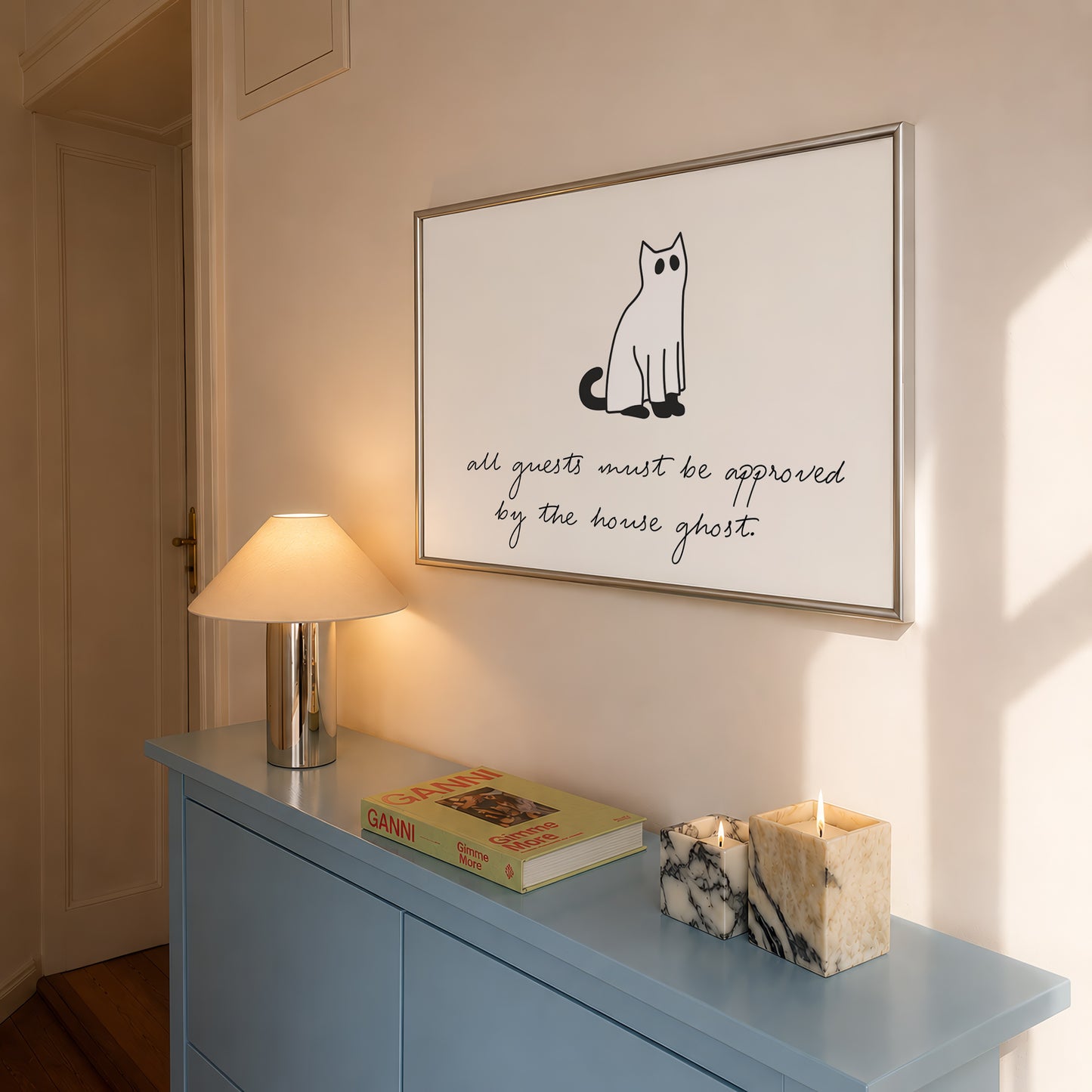 Horizontal minimalist Halloween print of a black cat wearing a white ghost sheet costume with cut-out eyes, above the handwritten cursive quote "all guests must be approved by the house ghost." on a cream background.