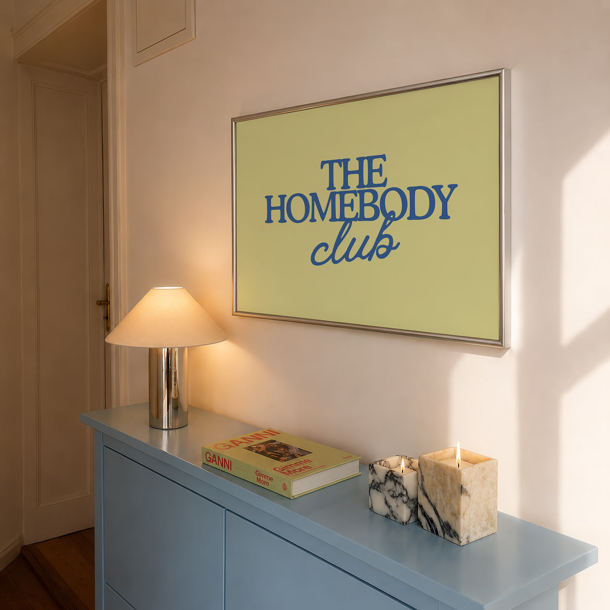 Rectangular landscape digital print with an olive-green background featuring "THE HOMEBODY" in bold blue serif type stacked above "club" in blue cursive script, centered typography layout.