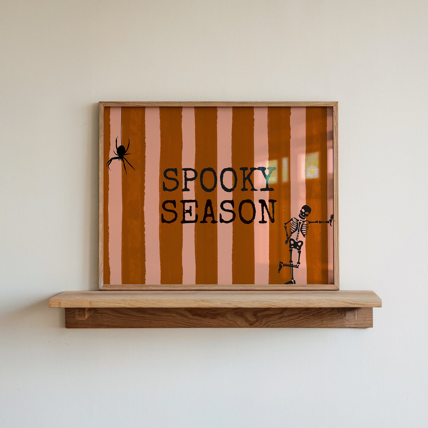 Horizontal print with orange and pink hand-painted vertical stripes, the words "SPOOKY SEASON" in bold black typewriter-style letters, a hanging black spider on the left, and a dancing skeleton illustration on the right.