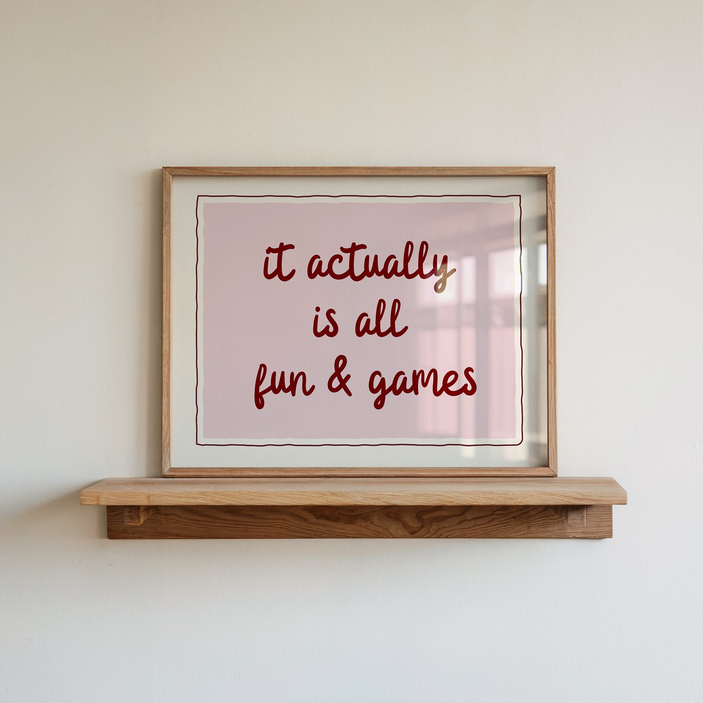 Horizontal wall print with a soft pink centre panel and white border with dark red scalloped outline, bold dark red cursive script reading it actually is all fun & games