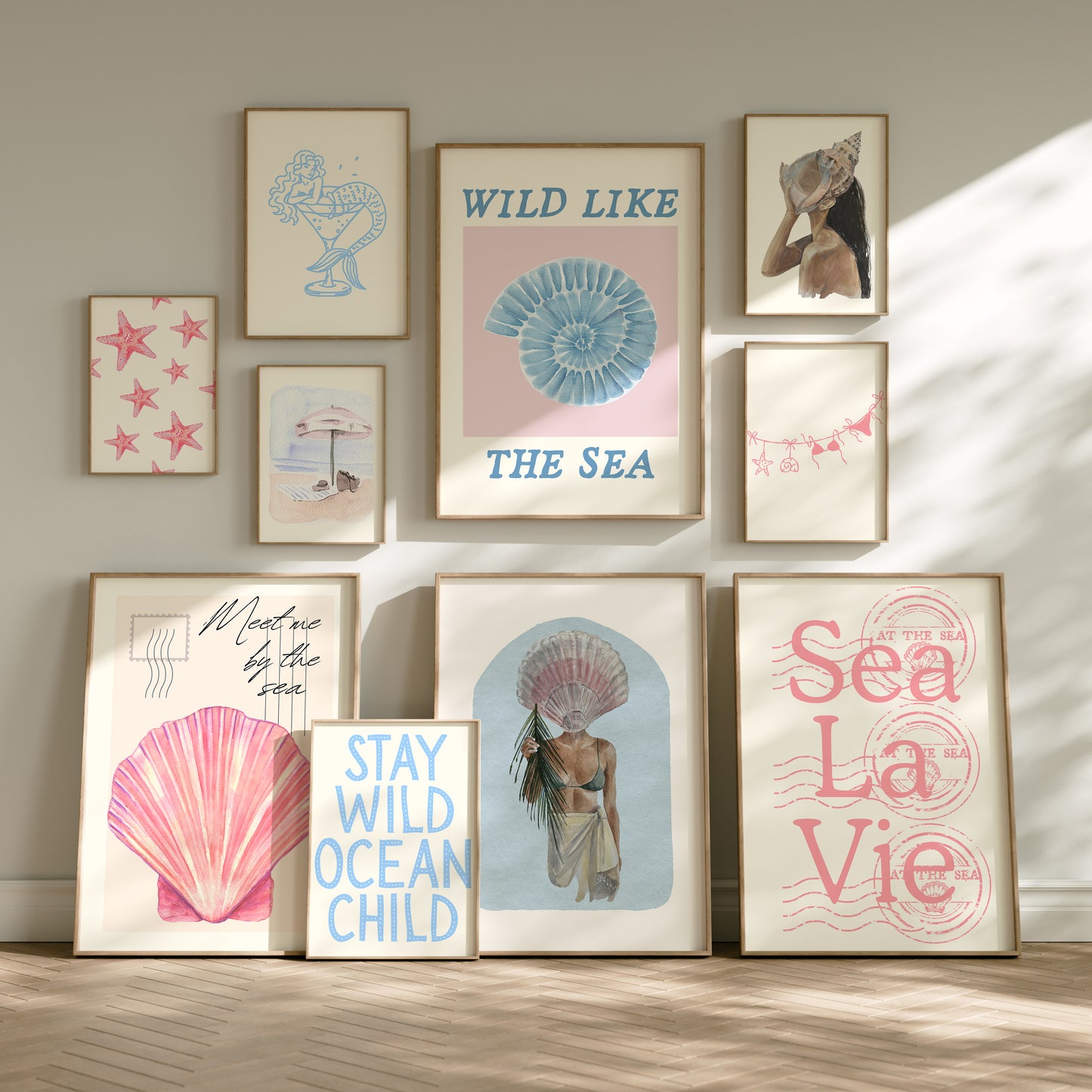 Set of 10 coastal watercolor prints in pink and blue: "Wild Like the Sea" beside a spiral shell illustration, "Stay Wild Ocean Child" in bold blue lettering, "Sea La Vie" in pink stamped typography, a mermaid lounging in a martini glass, pink starfish and seashell illustrations, a woman in a shell headdress, a beach umbrella watercolor, and "Meet me by the sea" in postcard-style script.