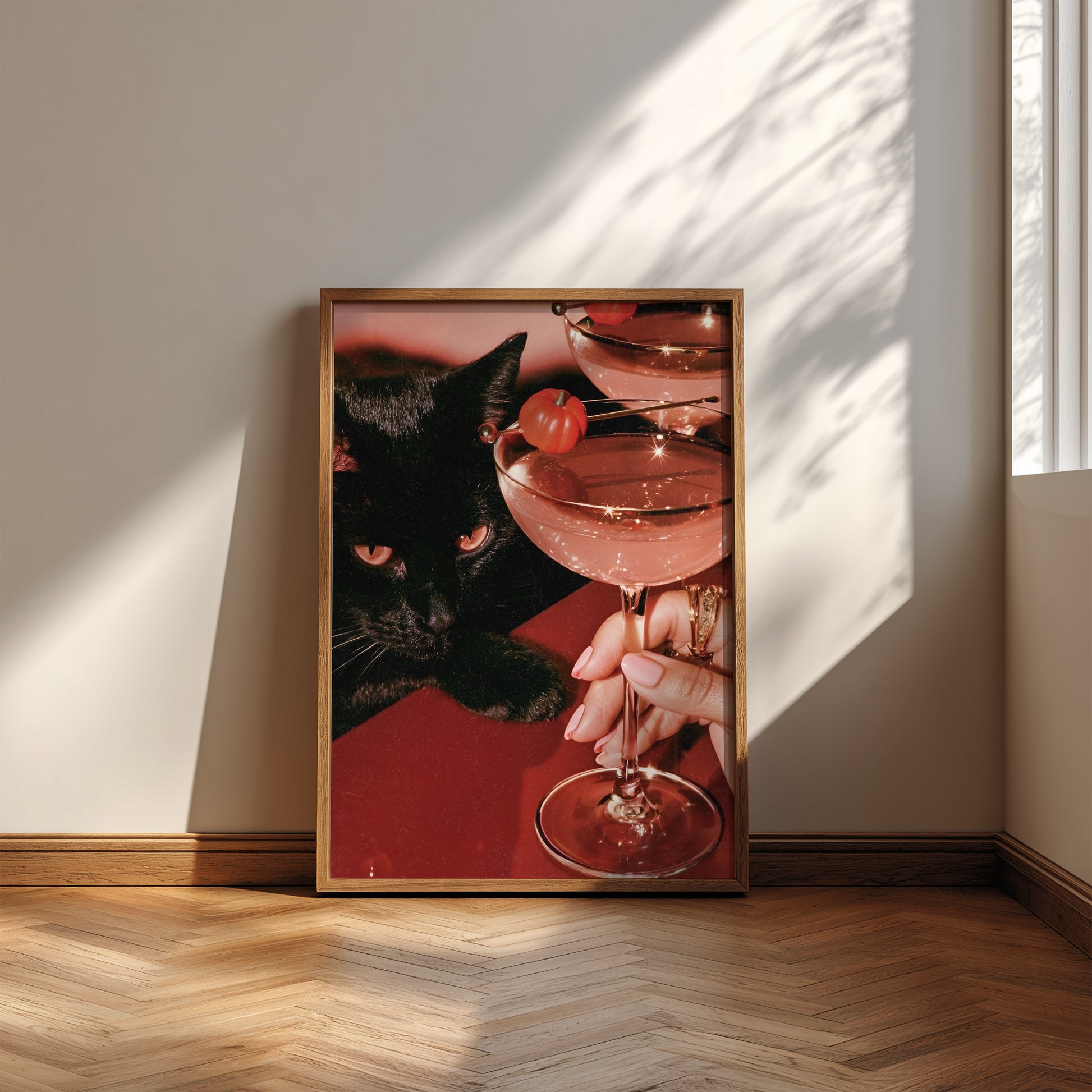 Vertical photographic print of a black cat with amber eyes resting beside two pink martini cocktails garnished with tiny orange pumpkins, on a deep red surface with a woman's hand holding one glass stem.