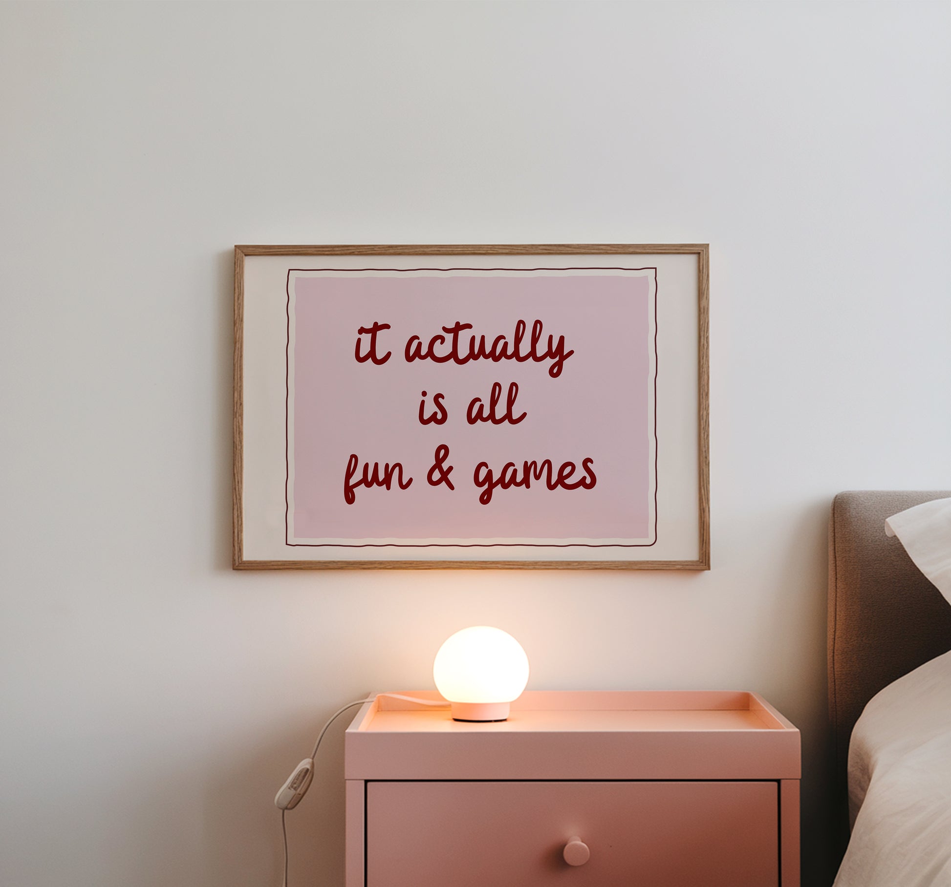 Horizontal wall print with a soft pink centre panel and white border with dark red scalloped outline, bold dark red cursive script reading it actually is all fun & games