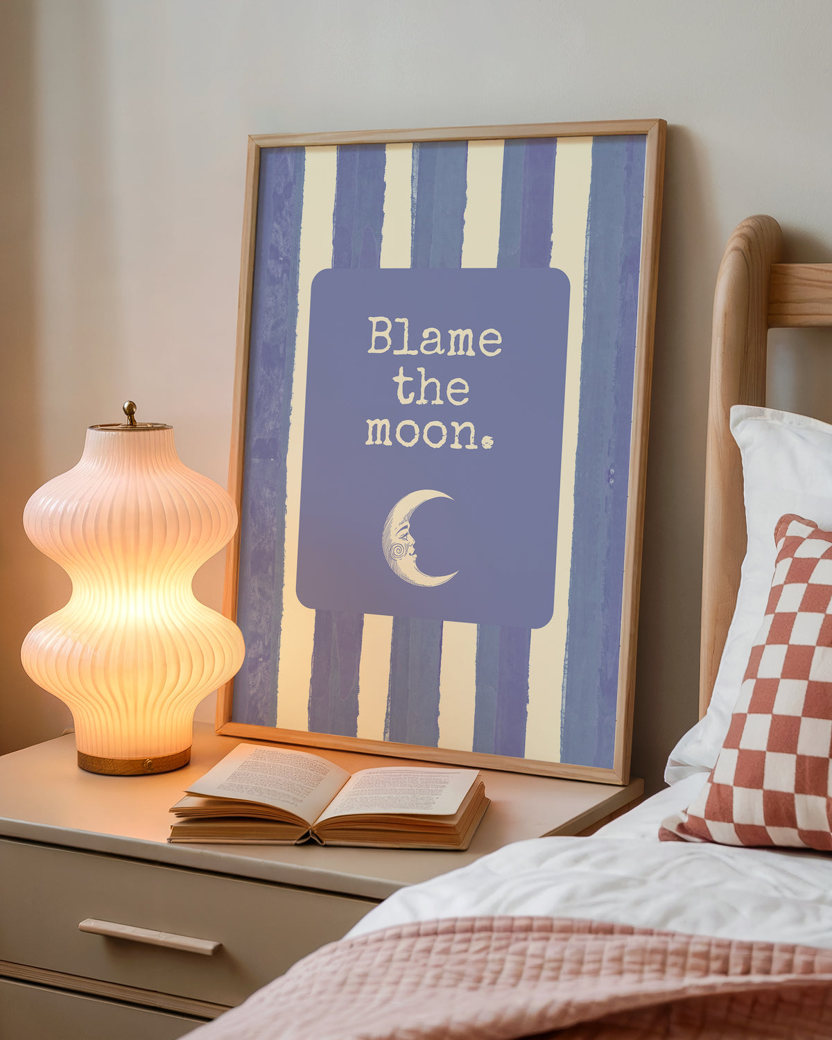 Portrait digital print with blue-and-cream striped background featuring a rounded blue panel with the text "Blame the moon." in cream lettering above a vintage-style crescent moon face illustration.