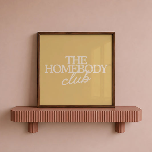 Square digital print with a mustard yellow background featuring "THE HOMEBODY" in bold cream serif type stacked above "club" in cream cursive script, centered typography layout.