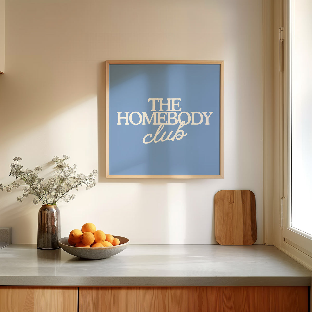 Square digital print with a dusty blue background featuring "THE HOMEBODY" in bold cream serif type stacked above "club" in cream cursive script, centered typography layout.
