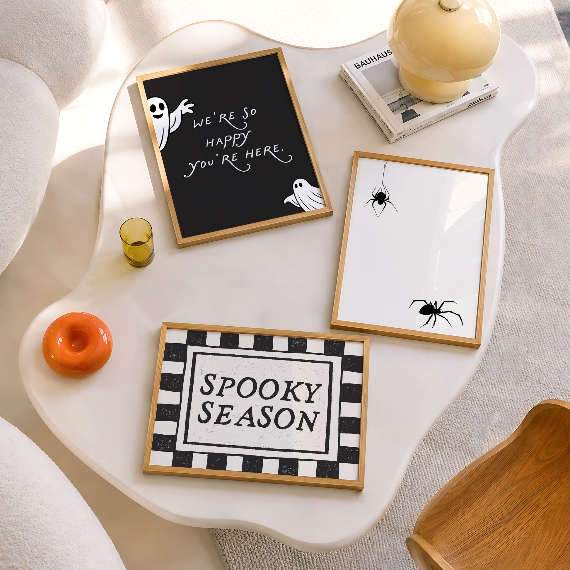 Set of 3 Halloween prints: a black background print with cartoon ghosts reading "We're so happy you're here," a checkered "Spooky Season" print, and a minimalist black spider illustration on a cream background.