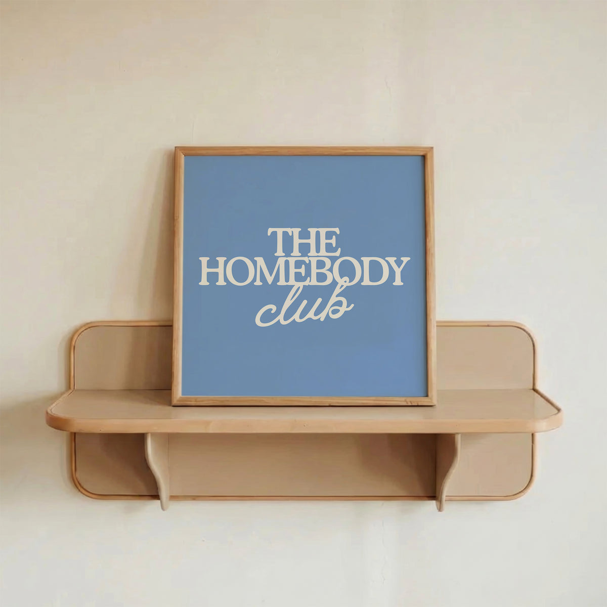 Square digital print with a dusty blue background featuring "THE HOMEBODY" in bold cream serif type stacked above "club" in cream cursive script, centered typography layout.