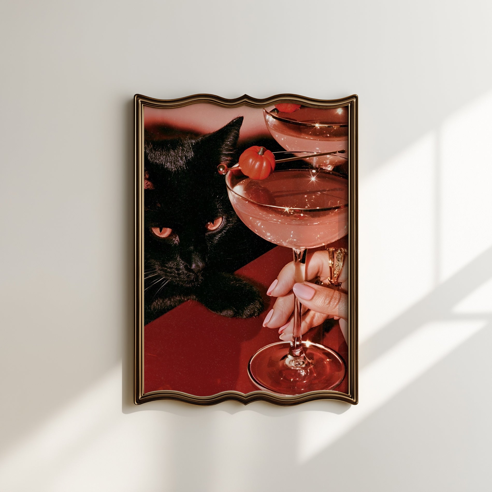 Vertical photographic print of a black cat with amber eyes resting beside two pink martini cocktails garnished with tiny orange pumpkins, on a deep red surface with a woman's hand holding one glass stem.