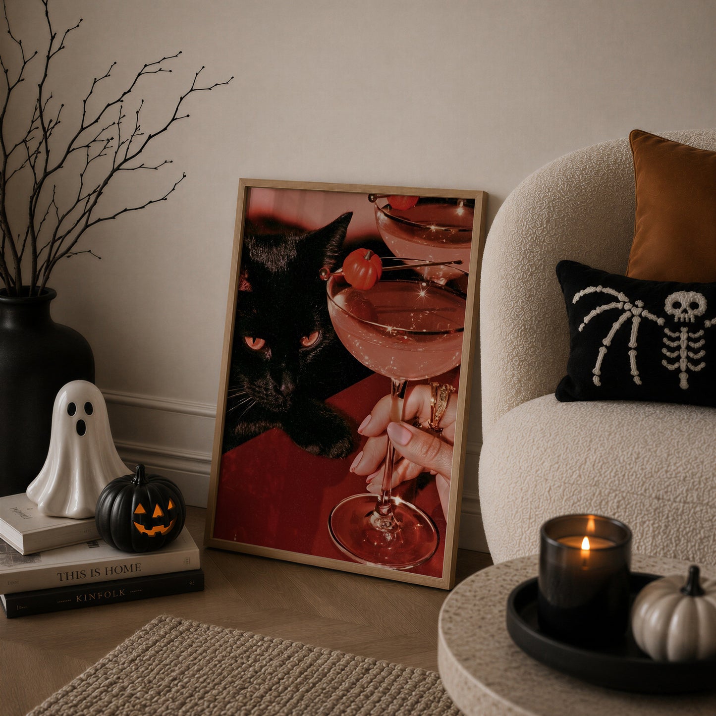Vertical photographic print of a black cat with amber eyes resting beside two pink martini cocktails garnished with tiny orange pumpkins, on a deep red surface with a woman's hand holding one glass stem.