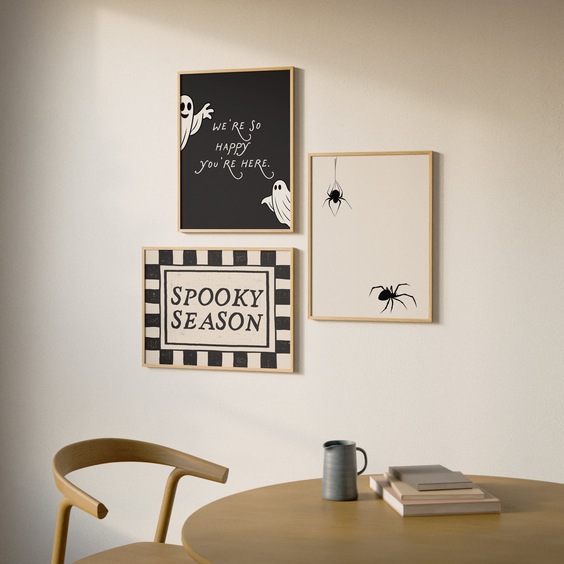 Set of 3 Halloween prints: a black background print with cartoon ghosts reading "We're so happy you're here," a checkered "Spooky Season" print, and a minimalist black spider illustration on a cream background.