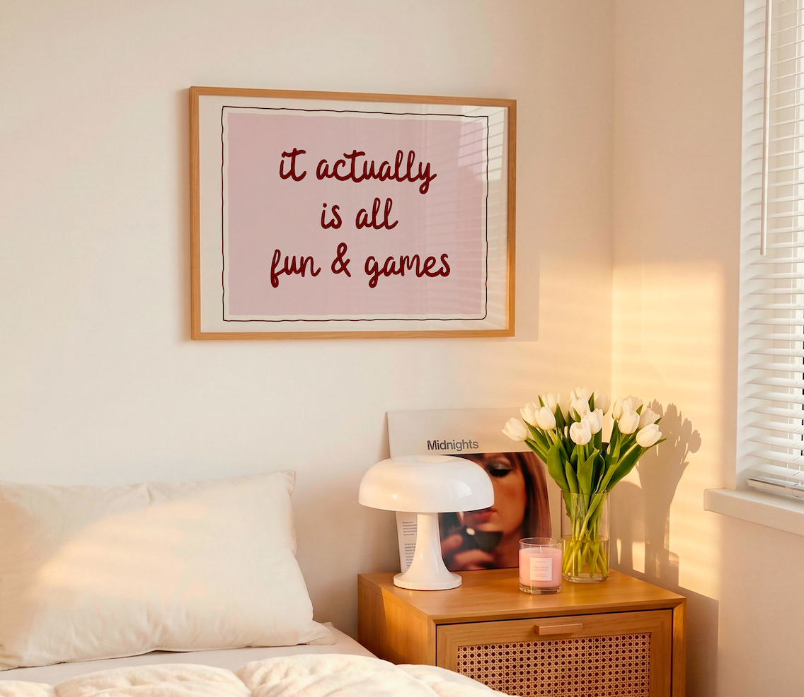 Horizontal wall print with a soft pink centre panel and white border with dark red scalloped outline, bold dark red cursive script reading it actually is all fun & games