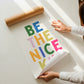 Vertical print with the words "BE THE NICE KID" stacked in large multicolored bubble letters on a cream background in a blue frame.